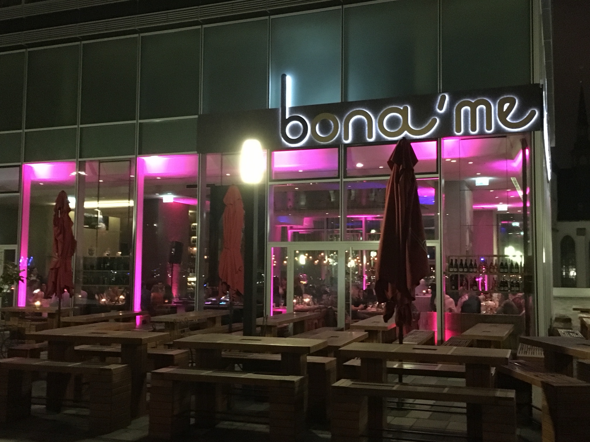 Außenansicht des Restaurants bona'me in Köln bei Nacht mit beleuchtetem Schriftzug und Außenterrasse