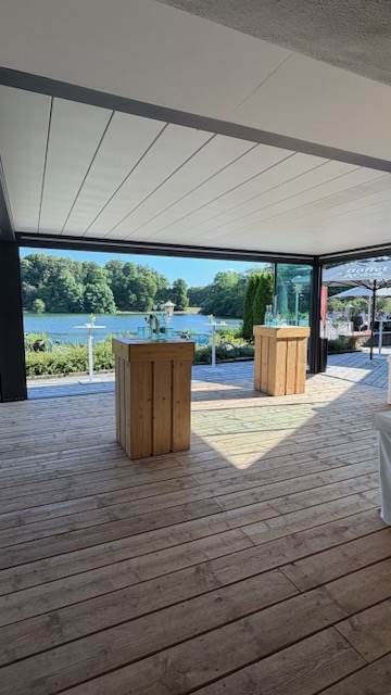 Überdachte Holzterrasse des Haus am See mit Stehtischen und Blick auf den Decksteiner Weiher